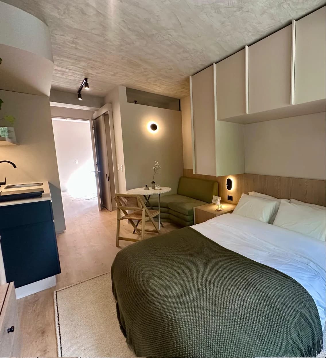 Apartaestudio con cama doble, cabecero de madera, sofá verde y cocina integrada